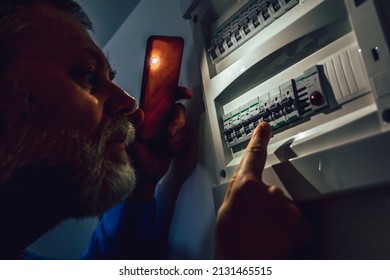 Energy crisis. Man in complete darkness holding a phone to investigate a home fuse box during a power outage. Blackout concept.