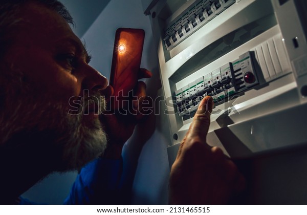 Energy crisis. Man in complete darkness holding\
a phone to investigate a home fuse box during a power outage.\
Blackout concept.