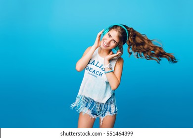 Energy girl with blue headphones  listening to music on blue background in studio. She wears white T-shirt, shorts. Long curly hair in tail is flying to side from move.