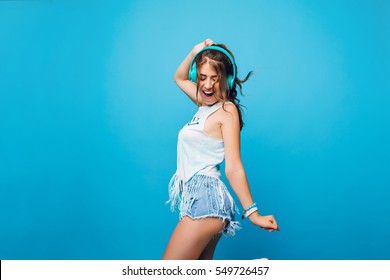 Energy girl with long curly hair in tail  listening to music with headphones on blue background in studio. She wears white T-shirt, shorts. She is dancing.