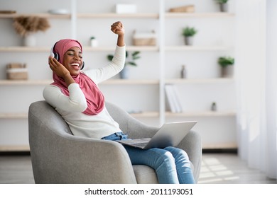 Energy Mood. Overjoyed Black Islamic Lady In Wireless Headphones Listening Music Online On Laptop While Relaxing In Comfortable Chair At Home, Cheerful Muslim Woman Raising Hand And Singing