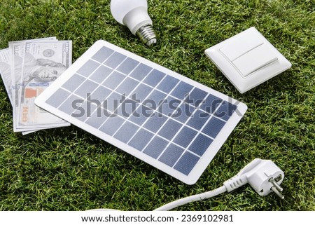 energy saving, power and sustainability concept - close up of solar battery model, lightbulb, money with plug and switch on green grass background
