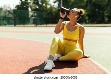 Energy, Vitality, Wellbeing Concept. Fit African American Woman In Wireless Earbuds Sitting On Basketball Field Ground, Holding Shaker, Drinking Water Or Protein, Taking Break After Outdoors Exercise