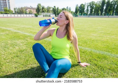 Energy, Vitality, Wellbeing Concept. Fit Sportswoman Sitting On Soccer Field, Holding Shaker, Drinking Water Or Protein, Taking Break After Outdoor Exercise.