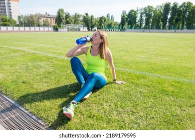 Energy, Vitality, Wellbeing Concept. Fit Sportswoman Sitting On Soccer Field, Holding Shaker, Drinking Water Or Protein, Taking Break After Outdoor Exercise.