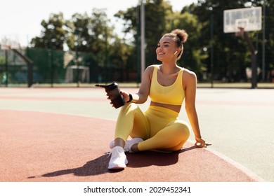 Energy, Vitality, Wellness Concept. Smiling African American woman sitting on basketball field ground, holding shaker, drinking water, taking break after outdoors exercise, wearing wireless earbuds - Shutterstock ID 2029452341