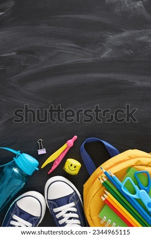 Engage with the educational process. Top view vertical photo of stationery for the school, child's rucksack, trendy shoes, water bottle on blackboard background with empty space for advert or text