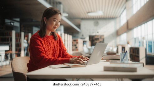 Engaged Asian Female Student Working on Her University Assignments With a Laptop in Modern Library. Young Woman in Red Sweater Studying, Surrounded by Books and Pens. - Powered by Shutterstock