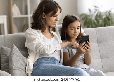 Engaged beautiful mom showing cute child girl how to use online application, service on mobile phone, resting on cozy soft home couch, touching smartphone screen, speaking, hugging kid - Powered by Shutterstock