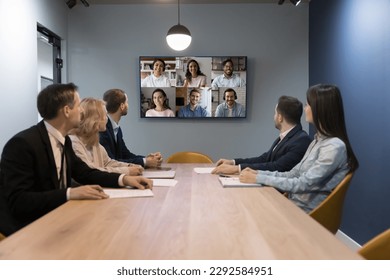 Engaged business team talking on online video conference call to multiethnic partners, sitting at large negotiation table in small office room, looking at display with diverse head shots - Powered by Shutterstock