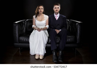 Engaged Couple Wearing Modern Wedding Suit And Bridal Dress In A Classical Retro Fashion Style.  The Stylish Groom And Bride Are Sitting On A Leather Couch In A Studio.