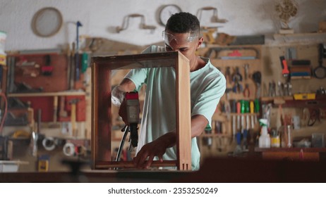 Engaged in Craft scene of a Young Black Brazilian Carpenter Building Furniture at Workshop, apprentice using drilling equipment, wood carpentry student - Powered by Shutterstock