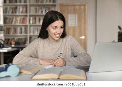 Engaged distant learning. Smiling young student girl enjoy productive self studying online sit at workplace with paper books on desk take written notes from video lecture webinar on notebook screen