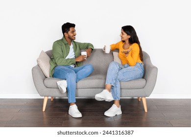 Engaged Indian spouses enjoying conversation with coffee mugs on cozy couch, in bright room with minimalist style and warm smiles, white wall - Powered by Shutterstock