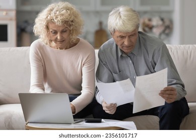 Engaged mature husband and wife reviewing paper bills, invoices, reading documents, using online payment app on laptop, paying medical insurance fees, doing domestic paperwork, calculating money - Powered by Shutterstock