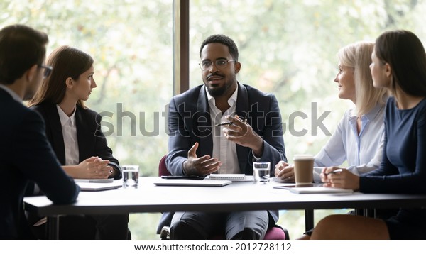 Engaged\
motivated African American business leader talking to team, meeting\
with employees, managers. Corporate coach, mentor training staff.\
Diverse group discussing work project at\
table