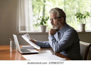 Engaged in paperwork. Serious aged businessman working from home office sit at desk read loan contract letter correspondence. Retired hoary man in glasses think on financial papers study bank report - Powered by Shutterstock