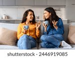 Engaged in a warm conversation, two young pretty women share stories over coffee on a beige couch at home
