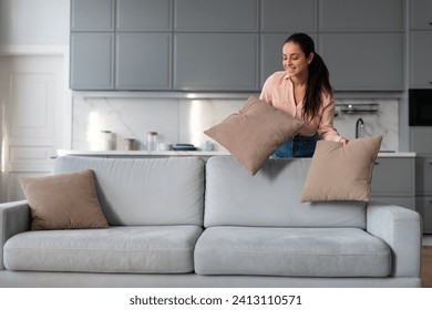 Engaged woman carefully arranging and fluffing throw pillows on sofa, taking pride in her neat and stylishly decorated living space with modern kitchen backdrop
