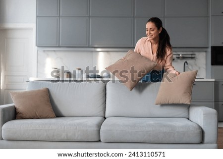 Engaged woman carefully arranging and fluffing throw pillows on sofa, taking pride in her neat and stylishly decorated living space with modern kitchen backdrop