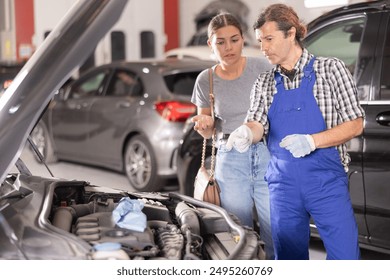 Engaged young female customer discussing necessary repairs or maintenance with experienced mature male mechanic next to open car hood in auto repair shop .. - Powered by Shutterstock