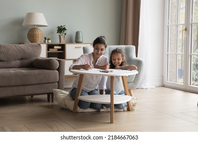 Engaged young Indian mother   happy little daughter kid drawing in pencils in living room  scratching doodles in paper album  sitting at small table  chatting  laughing  Family home leisure