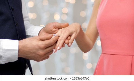 Engagement, Proposal And People Concept - Close Up Of Man Putting Diamond Ring On Womans Finger Over Holiday Lights Background