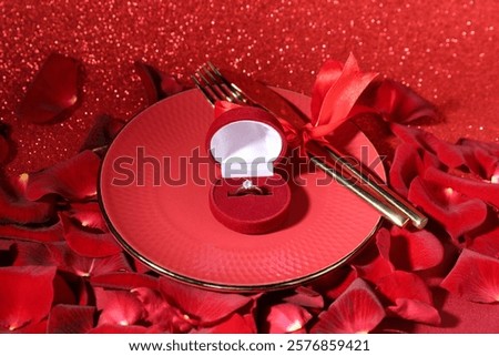 Engagement ring in box, tableware and rose petals on red glitter background, closeup. Table setting for romantic dinner