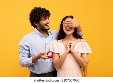 Engagement, romantic concept. Loving indian man making proposal with beautiful ring to his lovely girlfriend, closing lady's eyes with hand, standing over yellow background