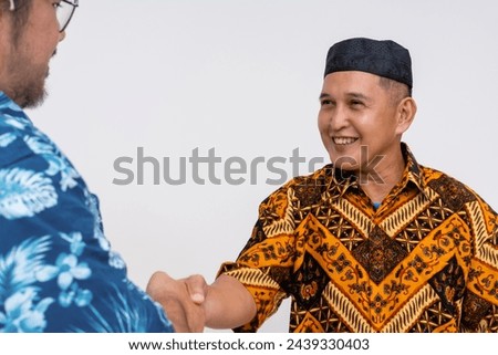 An engaging moment of cultural greeting, showing an Indonesian man in traditional batik and kopiah shaking hands with a tourist in a Hawaiian shirt.