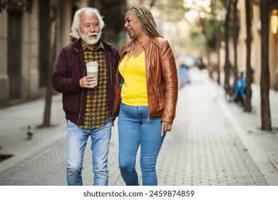 Engaging and relaxed senior couple sharing a laugh and a coffee while walking down a tree-lined urban street, enjoying each other's company. - Powered by Shutterstock