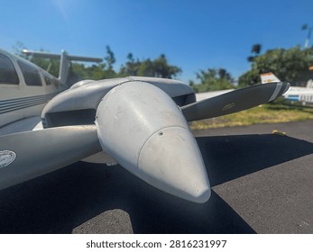 The engine nacelle and propeller of a twin-engine airplane parked on an asphalt surface. The airplane is white and has some blue stripes and a logo on its side. The propeller is a dark gray color. The background consists of green trees and a clear blue sky.