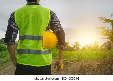 Engineer Or Architect, Wearing Green Vest, Holding Yellow Safety Helmet, Water Level Gauge Looking At Construction Site, Empty Land With Morning Sun Light Future Career, Home Building Construction 