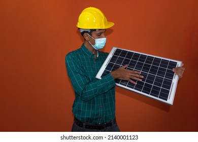 Engineer, Architect Or Worker With Yellow Helmet And Pandemic Mask Isolated On Orange Background. Engeneer At Work Holding A Photovoltaic Solar Panel.