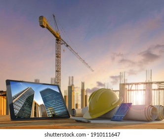 Engineer or architect's tools in construction site at sunset. Tablet, hard hat and blueprints on wooden table with crane and building rebars on the background. - Powered by Shutterstock