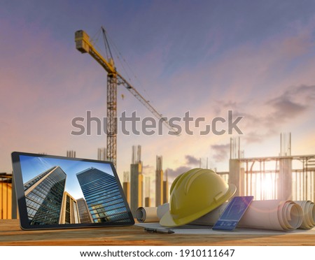 Engineer or architect's tools in construction site at sunset. Tablet, hard hat and blueprints on wooden table with crane and building rebars on the background.
