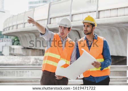 Engineer boss and foreman or worker working together at worksite in city. Employer guy pointing finger to construction for explaining project or plan. Blue collar worker hold blueprint, safety hats