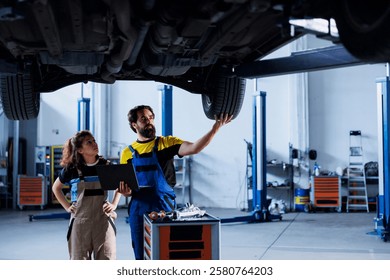Engineer in car service uses laptop to calculate invoice after fixing broken automobile wheels. Garage worker uses device to inform client of final costs after mending vehicle on overhead lift
