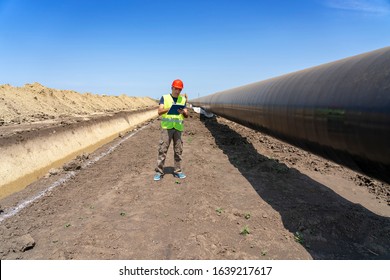 Engineer Checking Petrochemical Gas Pipeline At Construction Site - Turkish Stream. Portrait Of A Worker In Red Hard Hat And Yellow Vest. Engineer Standing Next To Oil Or Gas Pipeline.