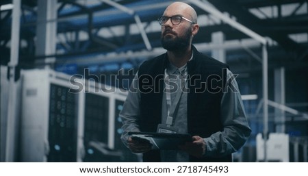 Engineer Checking System Metrics on Digital Tablet. Bearded IT Engineer in Dark Vest and Glasses Monitors System Metrics on Digital Tablet Amidst Server Racks. Cloud Auditing, and Technical Analysis.