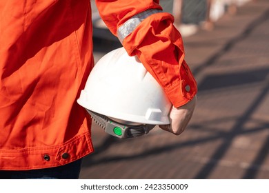 An engineer or construction worker is holding a white safety helmet, wearing orange coverall, standing on the working platform walkway. Ready to work in the challenge workplace concept scene. 