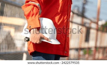 An engineer or construction worker is holding a white safety helmet, wearing orange coverall, standing on the working platform walkway. Ready to work in the challenge workplace concept scene. 