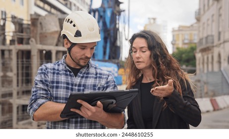 Engineer constructor discuss build plan with investor woman. Builder guy show construction site project tablet pad. Worker tell blueprint client. Man work city street. Customer look new house scheme. - Powered by Shutterstock