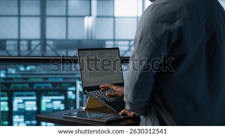 Engineer in data center operating notebook, deploying new server architectures. Server farm employee verifying settings using laptop, maintaining stable network performance, camera A