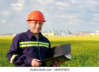 The Engineer Examines The Drawings Of The Plant For The Production Of Biodiesel On The Background Of Rapeseed Field