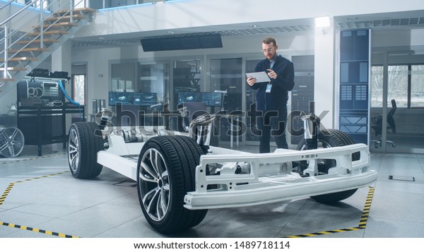 Engineer with Glasses and Beard Scans\
Electric Car Chassis Prototype with Wheels, Batteries and Engine\
with an Augmented Reality Software on a Tablet Computer in a High\
Tech Development\
Laboratory.
