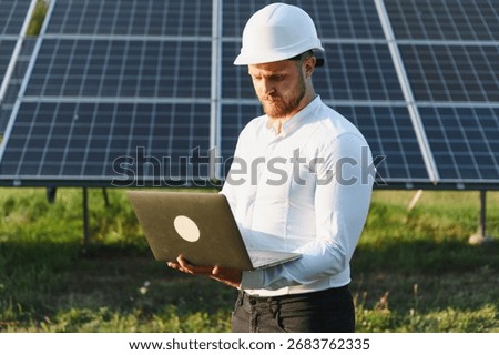 Engineer in hard hat checking solar photovoltaic panels with a laptop, monitoring sustainable renewable energy technology