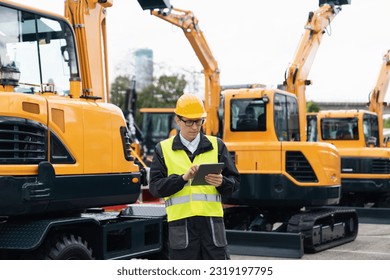 Engineer in a helmet with a digital tablet stands next to construction excavators	 - Shutterstock ID 2319197795