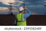 Engineer in helmet and safety vest past power transmission lines in semi dark field. Mature technician works night shift at power generation plant in country field. Electrician at power substation