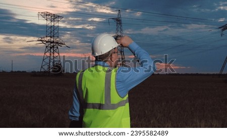 Engineer in helmet and safety vest past power transmission lines in semi dark field. Mature technician works night shift at power generation plant in country field. Electrician at power substation